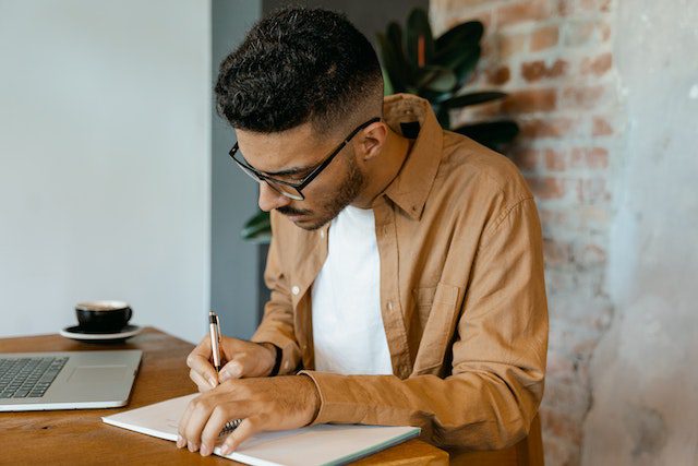 A Man Wearing a Brown Long Sleeve Shirt Writing on Notebook