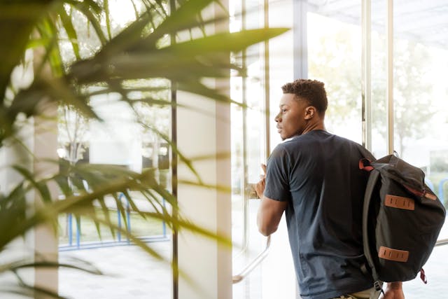 Man Looking Outside Window Carrying Black and Brown Backpack While Holding His Hand on Window