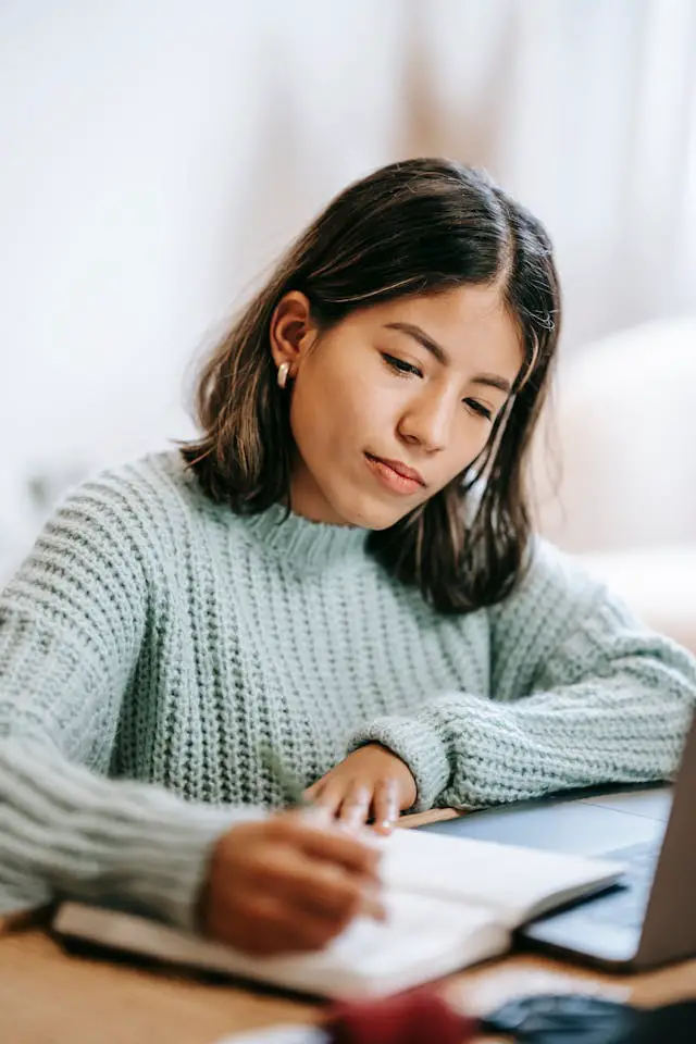 concentrated latin american lady writing information in notebook near netbook