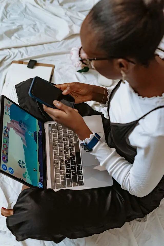 woman using smartphone and laptop while sitting on bed