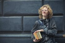 young man leaning against the wall holding a books