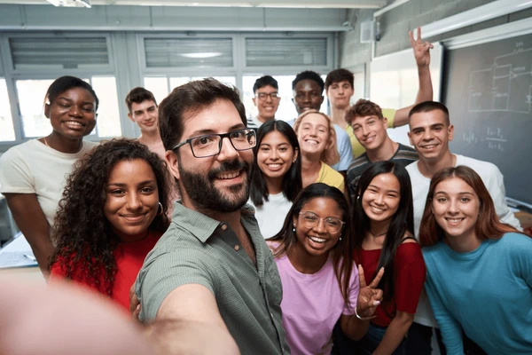 Happy vertical selfie of young group of students taking a photo with their male teacher in the classroom. Classmates from different countries,